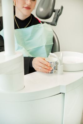 Patientin im Behandlungsstuhl haelt Becher neben Speibecken in einer Zahnarztpraxis