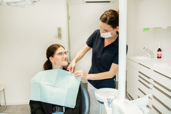 Zahnaerztin mit Mundschutz bereitet eine junge Patientin freundlich auf die Behandlung vor, beide befinden sich in einem modernen Behandlungszimmer.