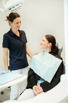 Eine Zahnarzthelferin steht in einem Behandlungszimmer während eine Patientin auf dem Behandlungsstuhl sitzt