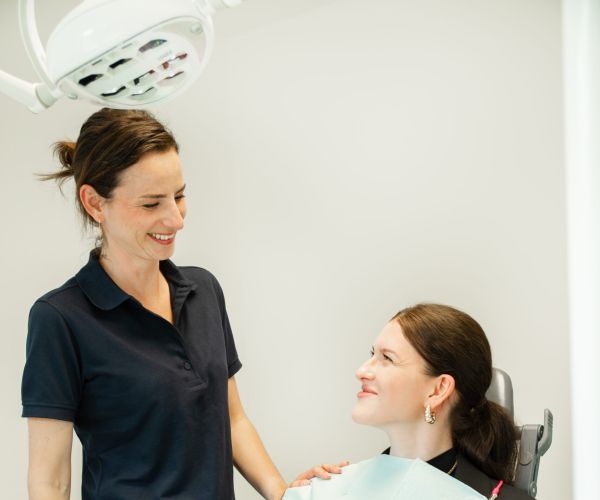 Eine Zahnarzthelferin steht in einem Behandlungszimmer während eine Patientin auf dem Behandlungsstuhl sitzt
